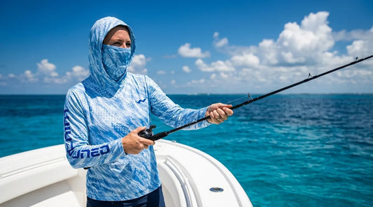 woman angler on a fishing boat in bright midday sun, wearing a hooded UPF fishing shirt, holding a rod over blue water, smiling confidently