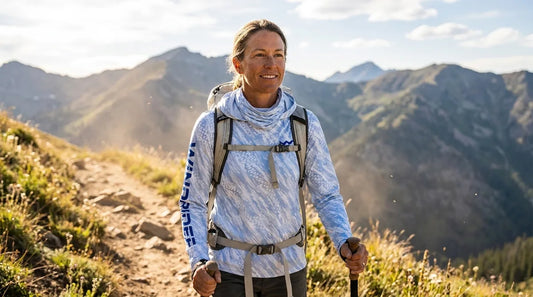 woman in her 40s hiking a sunlit mountain trail wearing a blue hooded long-sleeve sun shirt with hood down, trekking poles in hand, backpack straps visible across her chest