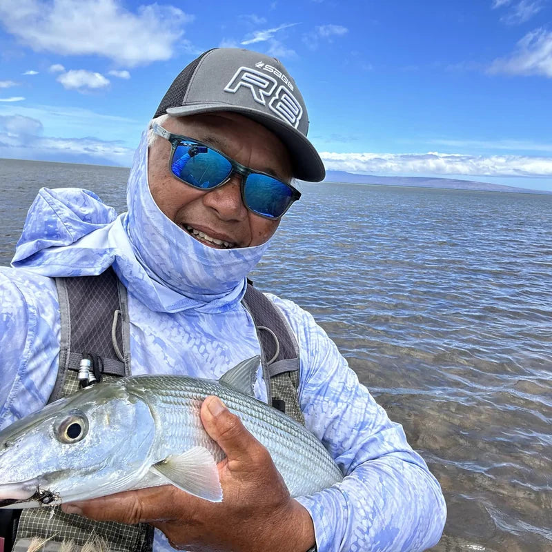 Angler on a flats boat in bright sun wearing a blue-camo Hooded Helios sun shirt with the hood up and gaiter pulled over his face, holding a bonefish above shallow turquoise water