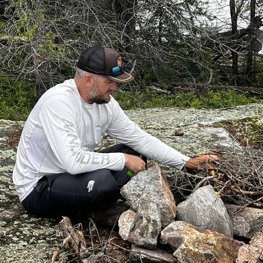 Man in white Helios sun shirt working outdoors building campfire on rocky shore
