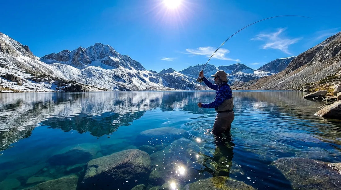 fly fisherman wading a high alpine lake, snowcapped peaks reflected in still water, midday sun high overhead, casting into deep blue mountain water