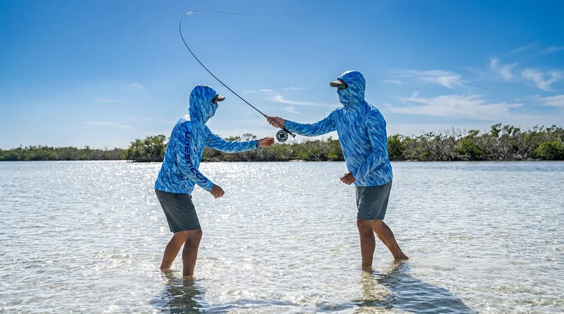 angler on bay flat in bright summer sun, wearing a hooded UPF shirt with integrated gaiter pulled up over nose and chin, rod in hand mid-cast, water reflecting sunlight