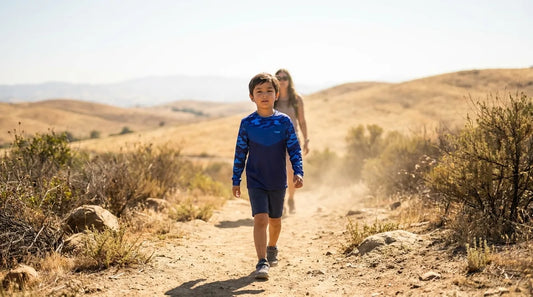 family hiking a dirt trail through open scrubland at midday, kid around age 6 in a long-sleeve sun shirt walking slightly ahead of a parent, dry hills and full sun in the background