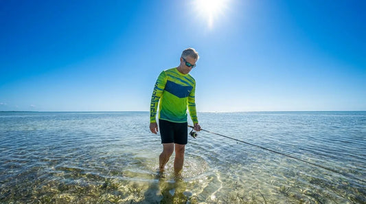 angler wade fishing shallow grass flats at midday, sun high overhead, staring down into clear water while holding a spinning rod, wearing a long-sleeve UPF shirt