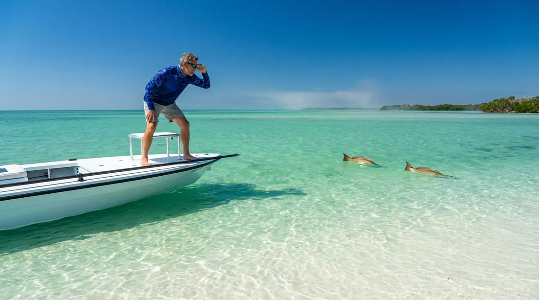 angler standing on the bow of a flats skiff in shallow Gulf Coast water, scanning for redfish tailing in the sun, wearing a long-sleeve UPF fishing shirt, bright midday light reflecting off white sand flat