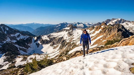 hiker above treeline in spring conditions, patchy snowfields visible, bright blue sky, strong midday light, wearing long-sleeve UPF sun shirt