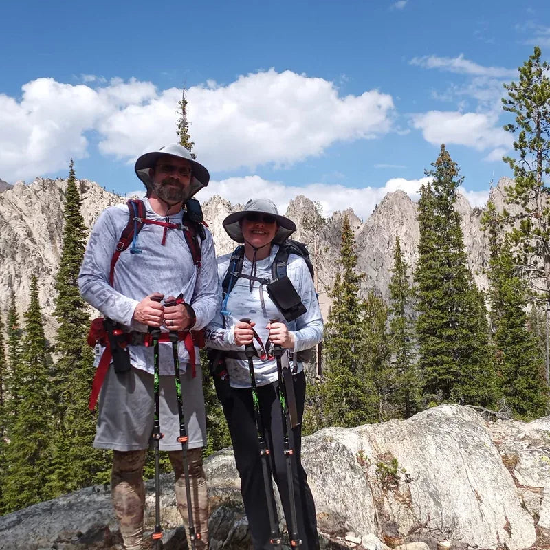 Couple at alpine summit wearing WindRider Helios hooded sun shirts with rocky mountain peaks behind