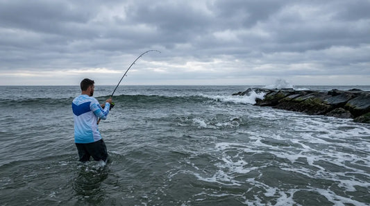 angler in long sleeve UPF shirt wading through surf toward breaking striper water at dawn, rocky jetty in background, overcast sky with silver light