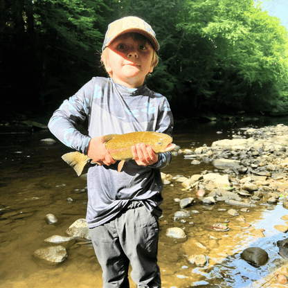 Young boy in grey HELIOS sun shirt holding trout by river wearing cap and sunglasses
